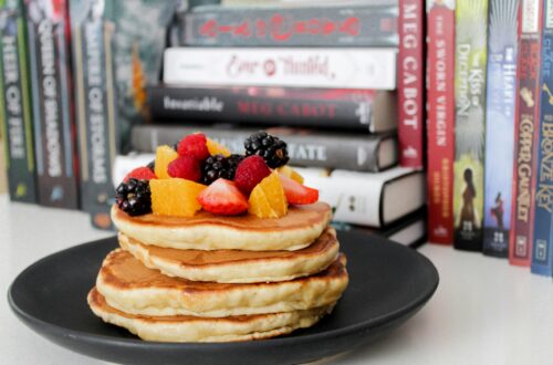 Stack of pancakes topped with fresh berries and oranges against a backdrop of books.
