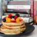Stack of pancakes topped with fresh berries and oranges against a backdrop of books.